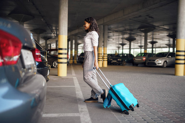 Giovane donna con valigia nel parcheggio auto. Viaggiatore donna con bagaglio nel parcheggio del veicolo, passeggero con borsa. Ragazza con bagaglio vicino automobile - Foto, immagini