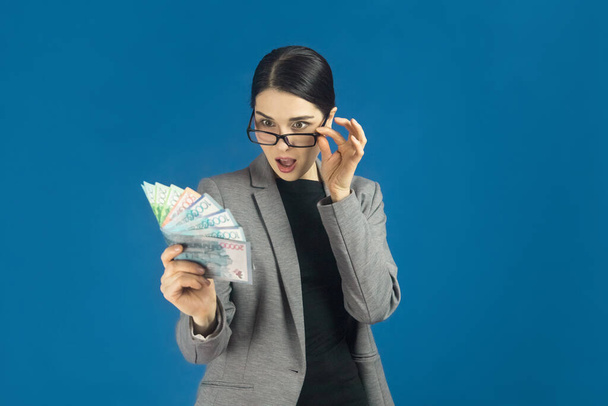Eine junge schöne Frau mit Brille schaut überrascht auf eine Packung Tenge in ihrer Hand. Selektiver Fokus - Foto, Bild