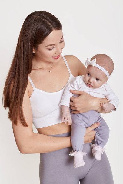 Smiling brown haired lady wearing bra holding new born girl with bow on her head, moomy looking at charming kid with love, posing isolated over white background. - Foto, Imagen