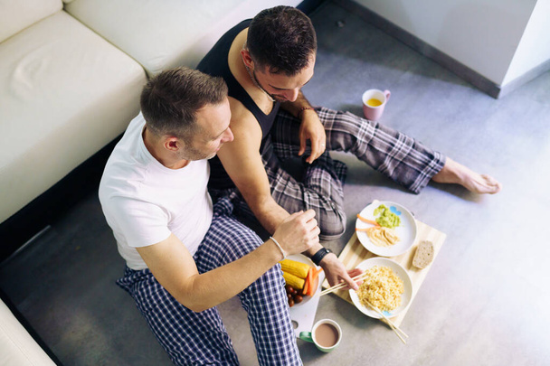 Gay couple eating together sitting on their living room floor. - Фото, изображение