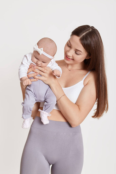 Winsome lady wearing bra and gray leggins, tiny cute infant with bow on head, smiling mommy with baby, posing isolated over white background, female with brown hair. - Foto, Imagen