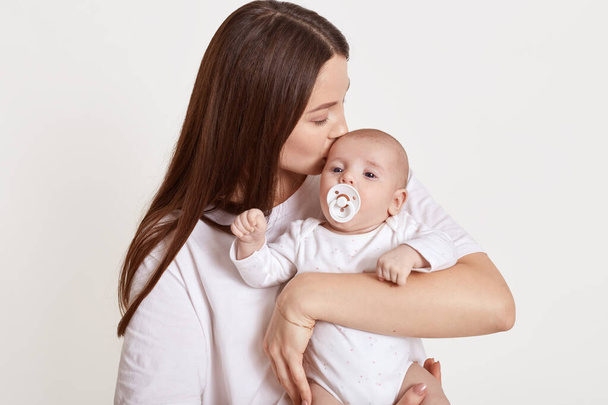 Mom with newborn baby posing against light background, mommy kissing her infant with nipple, attractive female feels happy, expressing gentle, wearing white clothing. - 写真・画像