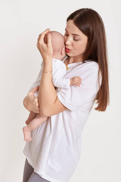 Brown haired mother dresses casual t shirt, holds her daughter in arms and kissing, posing isolated over white background, mommy with baby girl, love and care. - Zdjęcie, obraz