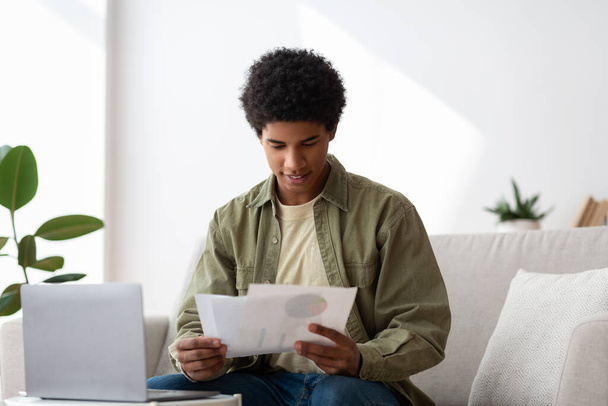 Happy black guy learning online from home, writing coursework, holding documents near laptop - Foto, afbeelding