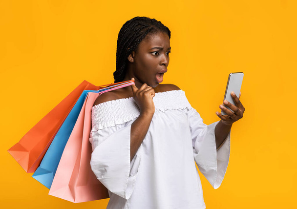 Shocked black shopaholic lady looking at smartphone and holding lots shopping bags - Zdjęcie, obraz