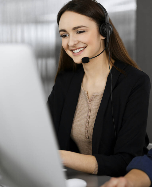 Young beautiful woman casual dressed in headsets is listening to a companys client, while she is sitting at the desk, working together with a male colleague in a modern office. Focus on woman. Call - Фото, зображення