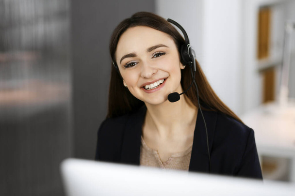 Young smiling girl in headsets is talking to the firms client, while sitting at the desk in a companys office. Portrait of a call center operator at work - Foto, Bild