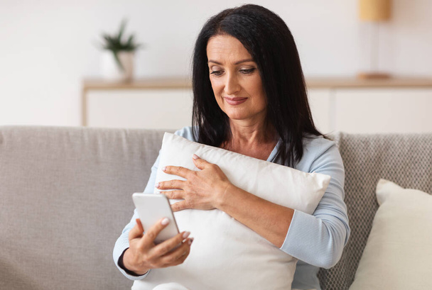 Smiling mature woman using mobile phone, holding pillow - Photo, image