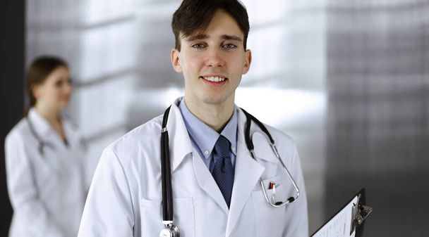 Friendly young male doctor standing with clipboard in modern clinic. Female colleague is busy with filling up of medication records form at the background of physician. Medicine concept - Photo, Image