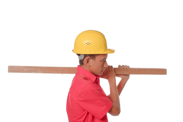 Bauarbeiter mit einem Holzbrett auf der Schulter. Tischler mit sefety Helm isoliert auf weißem Hintergrund. Schutzausstattungskonzept. - Foto, Bild
