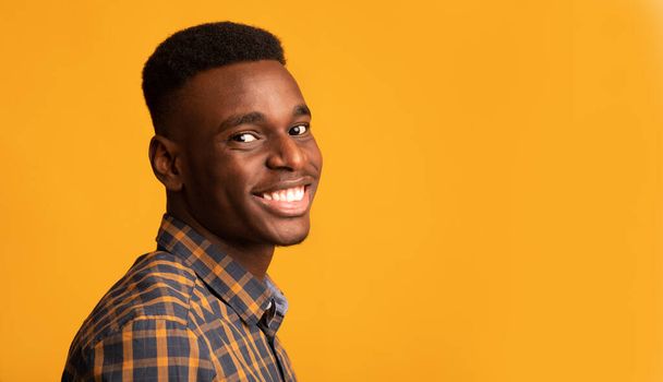 Mladý africký Američan se usmívá na kameru, stojící napůl obrácený přes žluté pozadí - Fotografie, Obrázek