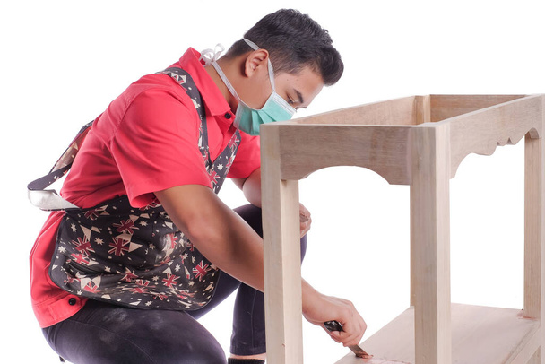 Jovem carpinteiro em roupas de trabalho usando raspador e enchimento de madeira no trabalho de acabamento para projeto de servidor de sofá isolado em fundo branco - Foto, Imagem