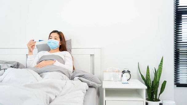 Chorą Azjatkę z maską ochronną leżącą w łóżku z wysoką gorączką i mierzącą temperaturę ciała za pomocą termometru i dotykającą czoła w domu - Zdjęcie, obraz