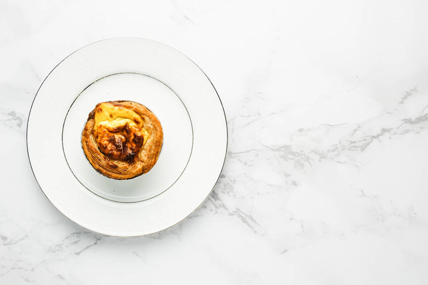 Flatlay of freshly baked egg tart on white plate on copyspace marble table. Famous Macao, Malaysia and Portugal snack food. - 写真・画像