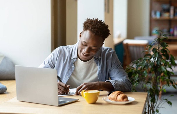 Happy black man studying remotely in front of laptop, taking notes during online class or lecture at city cafe - Photo, Image