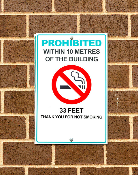 No smoking sign. Sign states that smoking is prohibited within 10 meters or 33 feet of the building. The wall it is on is brick. The sign appears to have been mounted over another sign.  - 写真・画像