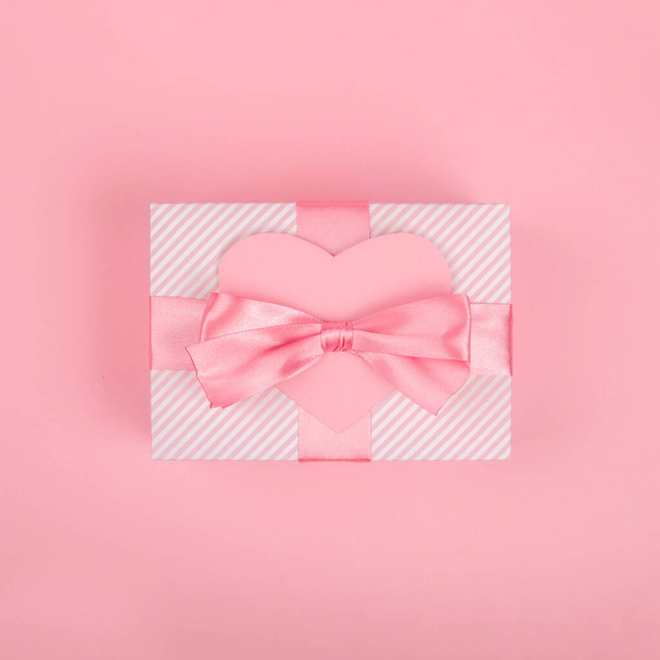 Valentine Day gift in a box wrapped in striped paper and tied with silk ribbon bow and heart shape greeting card on pink background with copy space for text - Foto, afbeelding