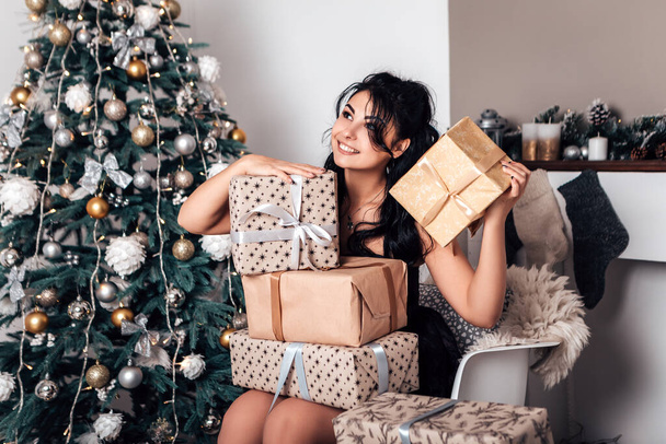 Brunette smiling happy woman opening wrapped ribbon gift box present, New Year evening, Christmas decorated tree. Lifestyle and shopping concept. Biracial girl greeting congratulating with holiday - Foto, imagen
