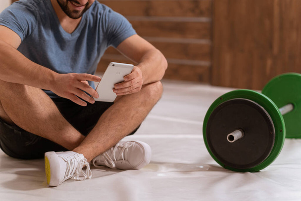 No face body shot. Young man searching online or texting sitting on a floor with smartphone, black and green tone fitness barbell, equipment for weight training concept. Healthy lifestyle concept - Foto, afbeelding