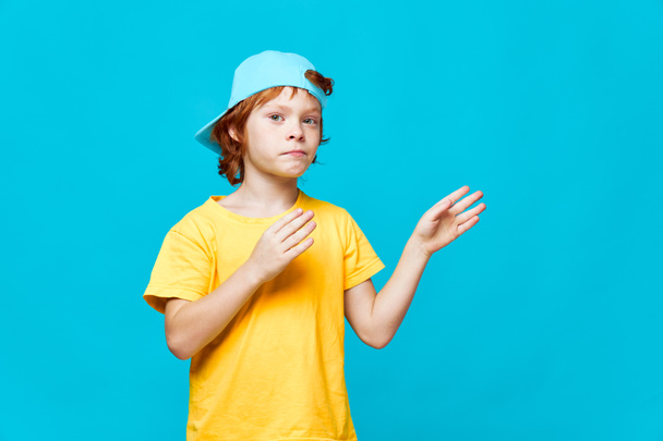 Red-haired boy gestures with his hands holding palms near him blue cap studio  - 写真・画像