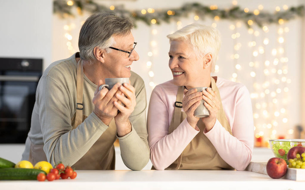 Ηλικιωμένο ζευγάρι γιορτάζει την παραμονή των Χριστουγέννων απολαμβάνοντας κακάο κάθεται στην κουζίνα - Φωτογραφία, εικόνα