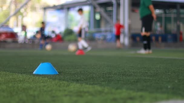 Marcador de cono azul y pelota de fútbol que se mueve sobre césped artificial verde con entrenamiento de equipo de fútbol infantil borroso en la academia de fútbol. - Metraje, vídeo
