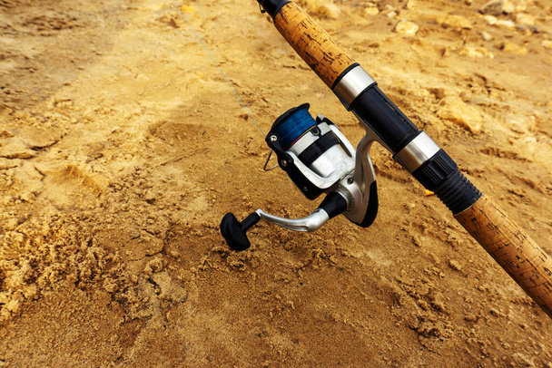 Summer fishing. A fragment of a spinning rod with a spinning reel on the background of a sandy shore. Copy space - Photo, image