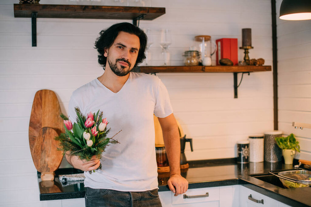Knappe man met een boeket in de keuken. Internationale Vrouwendag. Een jongeman houdt op 8 maart een boeket tulpen in zijn handen. Romantische familieavond thuis. - Foto, afbeelding