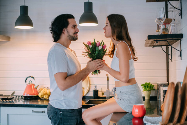 国際女性デー。自宅のキッチンで朝の若いカップル。男は美しいスタイリッシュなキッチンで3月8日にチューリップの花束を女性に与えます. - 写真・画像