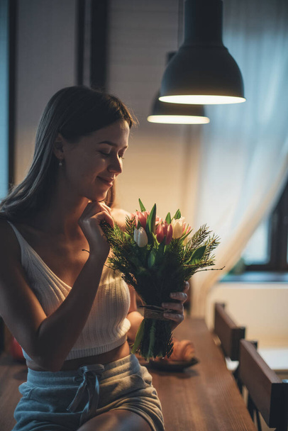 Portrét mladé ženy s kyticí květin. Krásná dívka sedí na stole pod večerním světlem lamp s tulipány v rukou. Smyslný portrét v tlumeném světle - Fotografie, Obrázek