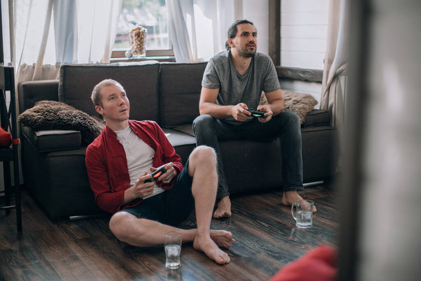 Kaverit pelaavat videopelejä sohvalla. Kaksi nuorta miestä, joilla on ohjaussauvat käsissään, pelaavat innokkaasti peliä konsolilla. - Valokuva, kuva