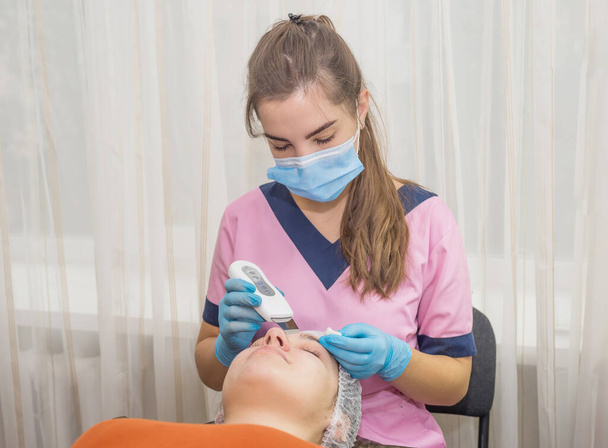chica cosmetóloga hace el procedimiento para la limpieza ultrasónica de la cara a su paciente en el consultorio de cosmetología - Foto, Imagen