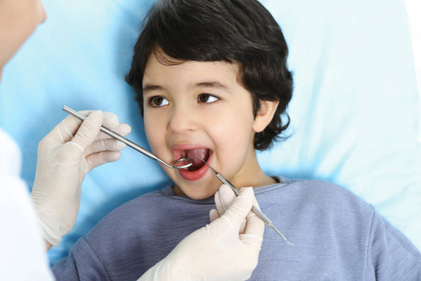 Cute arab boy sitting at dental chair with open mouth during oral checking up with doctor. Visiting dentist office. Stomatology concept - Foto, Bild