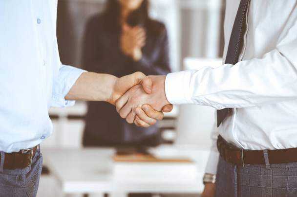 Two businessmen are shaking hands in office, close-up. Happy and excited business woman stands with raising hands at the background. Business people concept - Foto, imagen