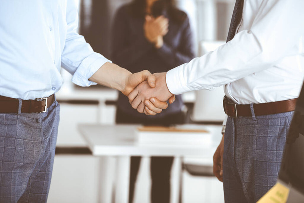 Two businessmen are shaking hands in office, close-up. Happy and excited business woman stands with raising hands at the background. Business people concept - Фото, зображення