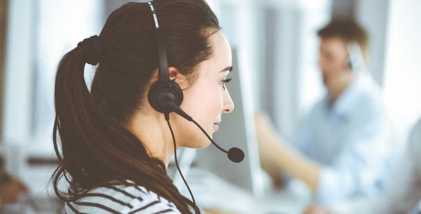 Business woman using headset for communication and consulting people at customer service office. Call center. Group of operators at work at the background. Casual dress style - Zdjęcie, obraz