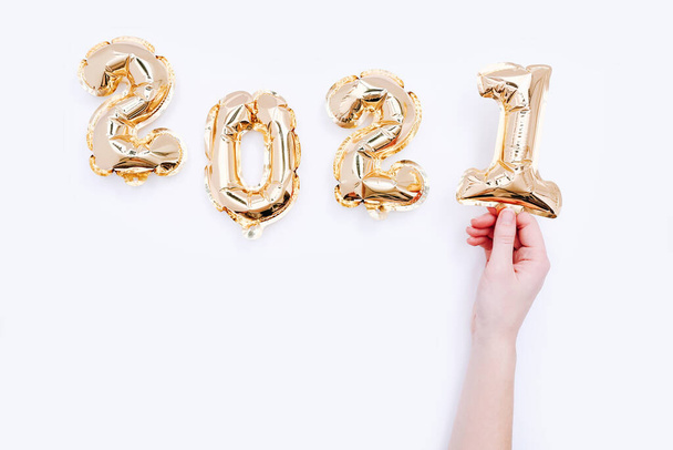 Globos de papel de aluminio en forma de números 2021 y mano femenina sosteniendo el número uno sobre fondo blanco. Celebración del año nuevo. - Foto, Imagen