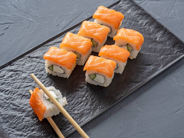 O sushi de Filadélfia rola em um suporte de placa texturizado preto em um fundo cinza. Um rolo é tomado em paus de bambu. Vista lateral, comida japonesa - Foto, Imagem