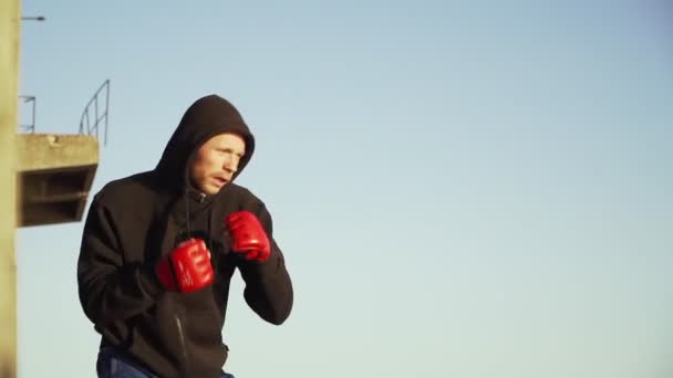 Caucásico Hombre boxeador en guantes rojos en una sudadera negra con capucha hace un ejercicio de lucha sombra - Metraje, vídeo