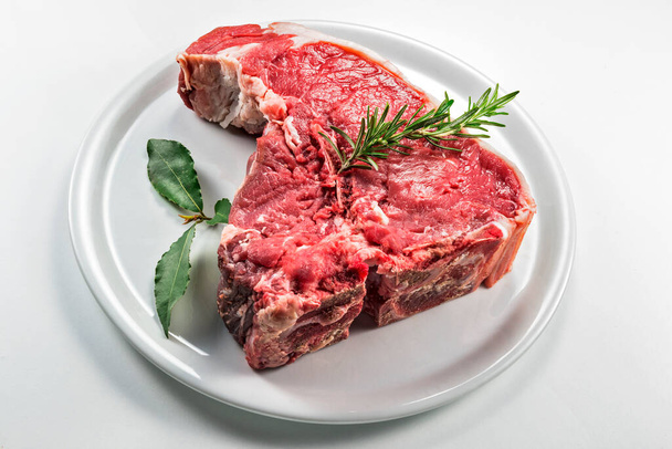 Raw T-bone steak with bay leaf and rosemary on white round plate - Photo, Image