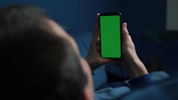 Hombre acostado en el sofá usando un teléfono inteligente con pantalla verde croma por la noche, desplazándose a través de las redes sociales o tienda en línea Internet, concepto de comunicaciones de cerca. - Metraje, vídeo
