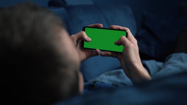 Hombre acostado en el sofá usando un teléfono inteligente con pantalla verde croma por la noche, desplazándose a través de las redes sociales o tienda en línea Internet, concepto de comunicaciones de cerca. - Metraje, vídeo