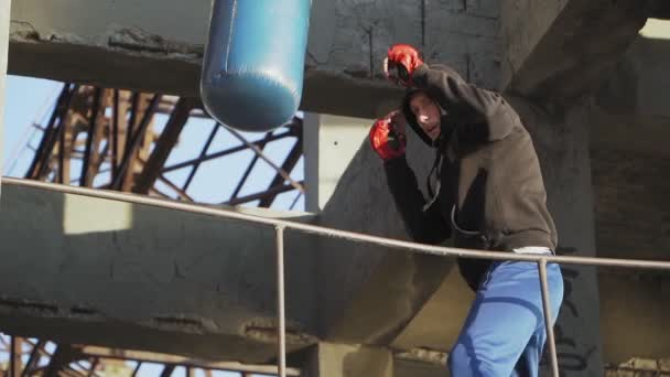 Boxer garçon caucasien frappe sac de frappe en arrière-plan un bâtiment abandonné. Boxe et arts martiaux - Séquence, vidéo