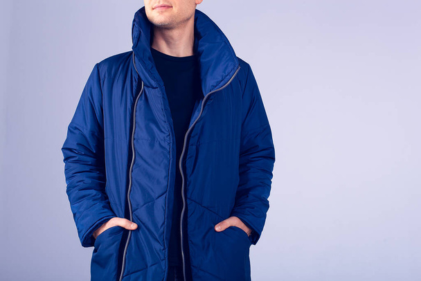 Модель парня на сером фоне одета в синюю куртку. Руки в карманы теплой куртки. Парень с бородой и тепло одетый. Стильная теплая одежда - Фото, изображение