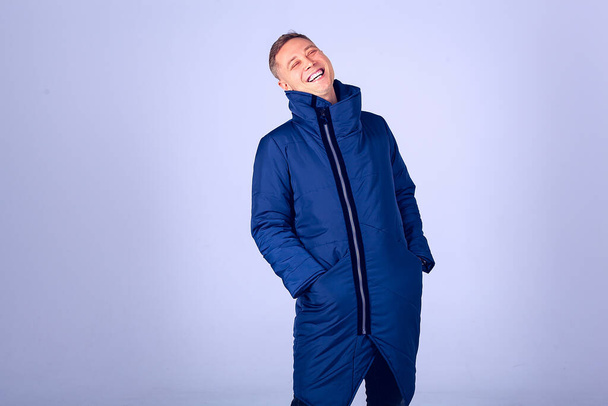 The man laughs hiding his hands in the pocket of a warm jacket. The body is bent with laughter. The guy on a gray background in a dark blue down jacket - Foto, afbeelding