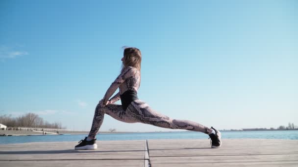 Η γυναίκα κάνει ασκήσεις στο φόντο μιας γαλάζιας λίμνης. Κάνοντας αθλήματα και προπόνηση φυσικής κατάστασης υπαίθρια - Πλάνα, βίντεο