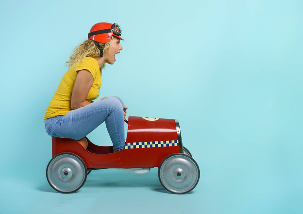 Vrouw met rode helm rijdt in een snelle speelgoedauto. cyaan achtergrond. - Foto, afbeelding