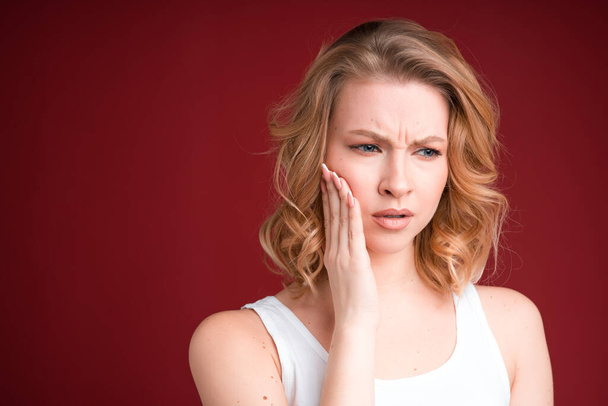 Blond woman with curly hair looks puzzled or having toothache in white tank top on red background. - Photo, Image