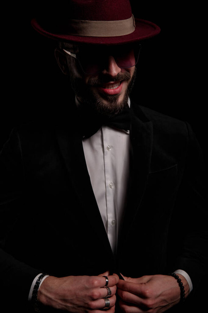 homme d'affaires heureux avec chapeau et lunettes de soleil souriant sur fond noir en studio, regardant de côté et fixant smoking velours noir - Photo, image