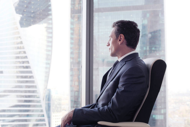 Business man sitting on armchair thinking looking at city view in modern glass office - Фото, зображення